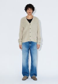 Beiger, übergroßer Cardigan aus flauschigem Material mit großen Knöpfen, kombiniert mit hellblauen, weit geschnittenen Jeans und grauen Schuhen. Weißer Hintergrund.