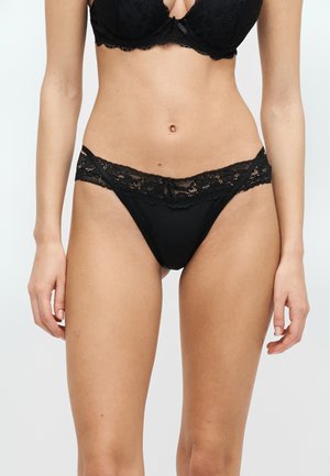 Μαύρο δαντελένιο σουτιέν και ταιριαστό σλιπ με ψηλή εφαρμογή δαντέλας, φορεμένα από έναν λεπτό άτομο σε ουδέτερο φόντο.