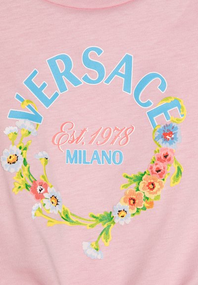 Ljust rosa bomullströja med blå och rosa text "VERSACE Est. 1978 MILANO" omgiven av ett blommönster.