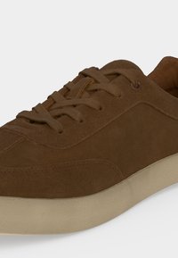Baskets en daim marron avec un design lacé et une semelle en caoutchouc beige, présentées en gros plan de côté.