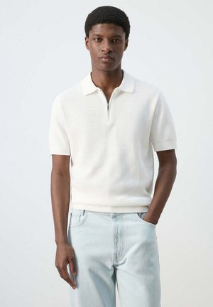 Joven con una camisa polo de punto blanca de manga corta con cremallera, vaqueros azul claro, de pie con una mano en el bolsillo.