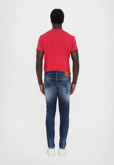 Camiseta roja de manga corta combinada con pantalones vaqueros azules de lavado desvanecido, con un ligero desgaste y bolsillos traseros con remaches. Zapatillas blancas.