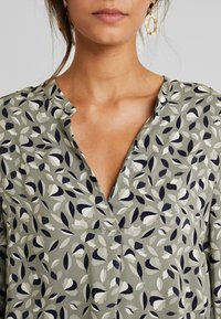 Femme portant un blouse verte avec des motifs de feuilles blancs, noirs et beiges et de grandes boucles d'oreilles ovales texturées en or, montrée de la nuque au buste.