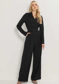 Combinaison noire à manches longues, boutons double boutonnage, design ajusté, jambes larges et poches latérales. Tissu de texture lisse.