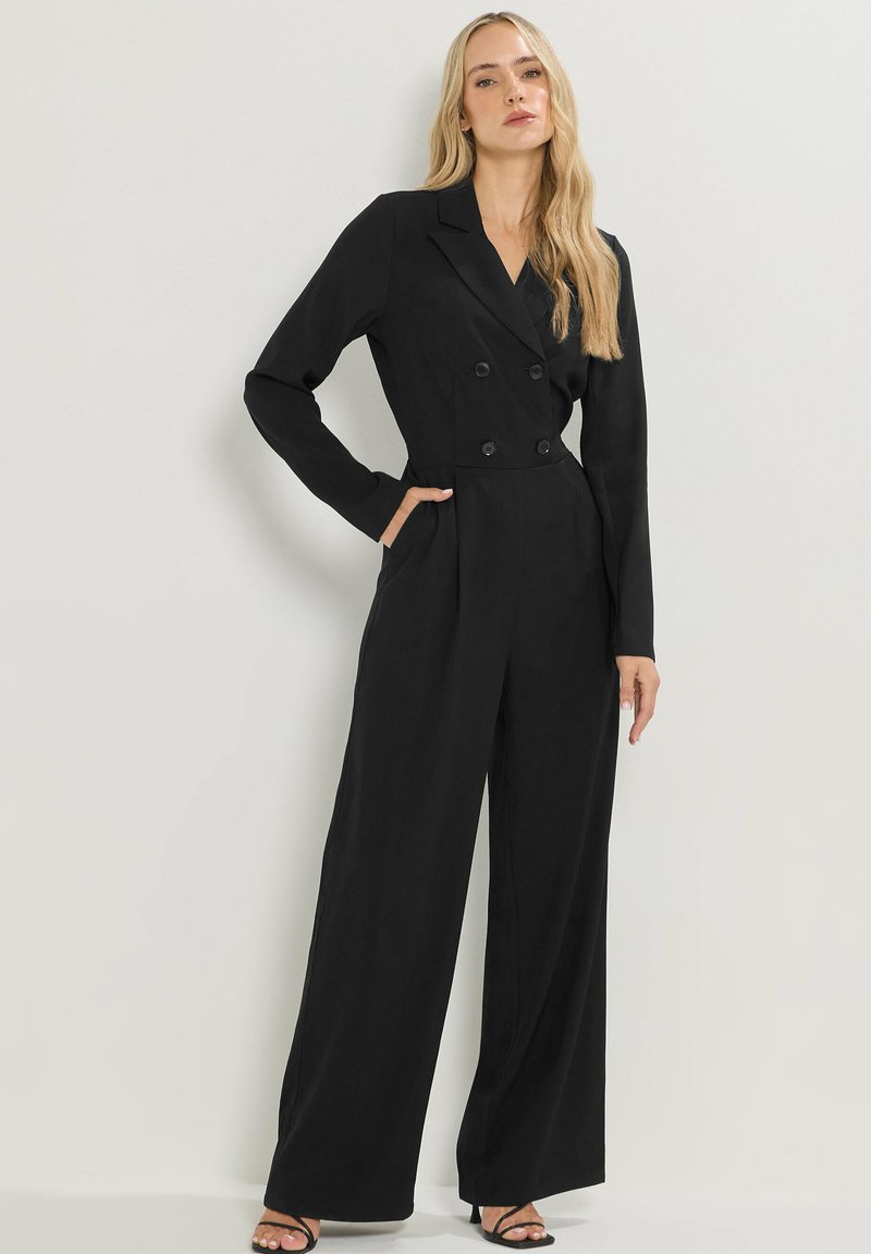 Combinaison noire à manches longues, boutons double boutonnage, design ajusté, jambes larges et poches latérales. Tissu de texture lisse.