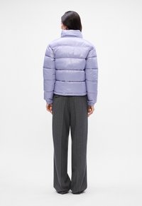 Chaqueta acolchada de color lavanda con un cuello alto, que presenta costuras horizontales. Se lleva sobre pantalones de pierna ancha de rayas gris, vista desde la parte de atrás.