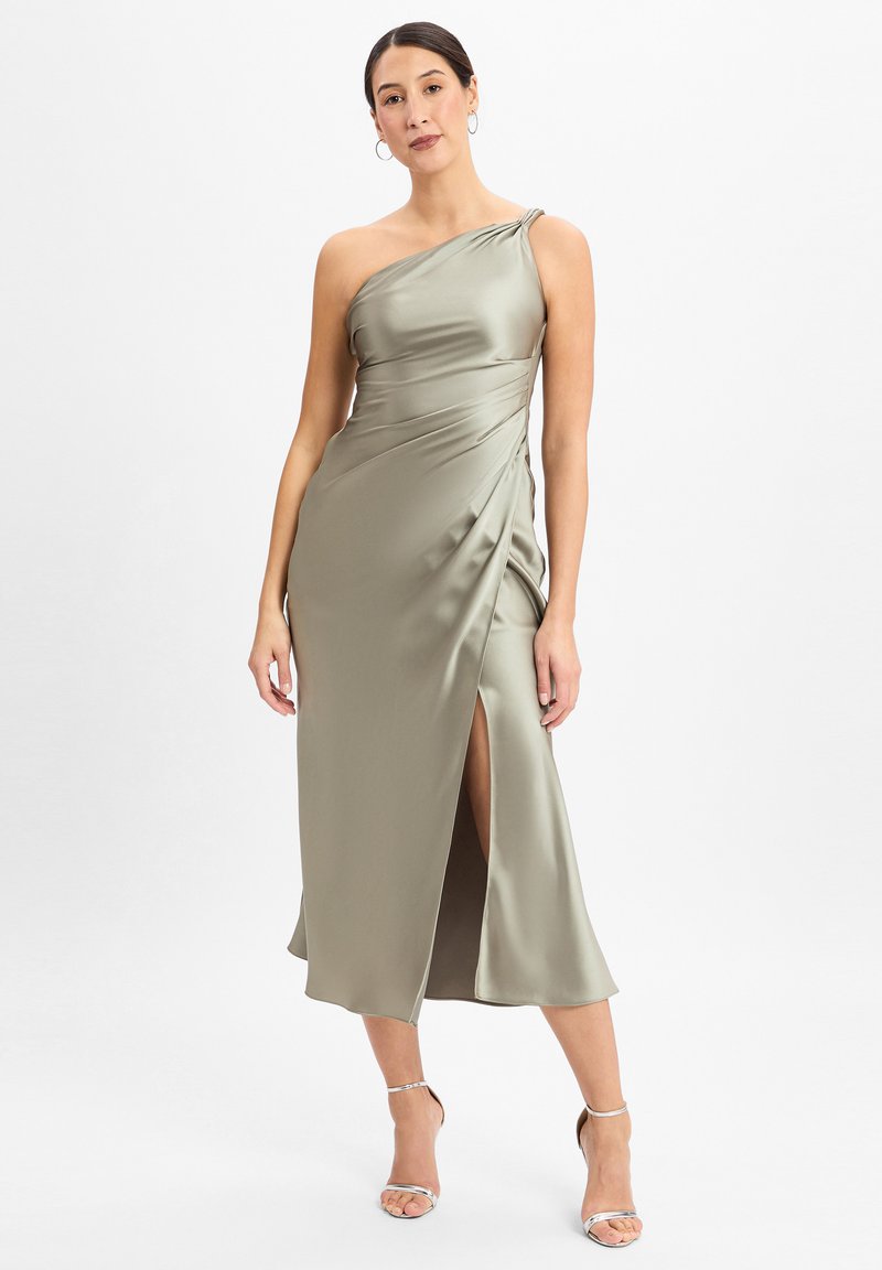 Satin, olijfgroen, één-schouder jurk met gedrapeerd ontwerp en zijsplit. Model in zilveren sandalen met bandjes, staand tegen een neutrale achtergrond.