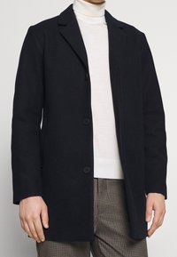 Marineblauer Mantel aus Wollmischung, einreihiges Design mit zwei Knöpfen, fallenden Revers und gerade geschnittenen Ärmeln. Kombiniert mit einem leichten Pullover.