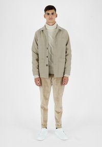 Giacca di lana beige con due tasche frontali, indossata sopra un maglione color crema a trecce, abbinata a pantaloni in velluto a coste marrone chiaro e sneakers bianche.
