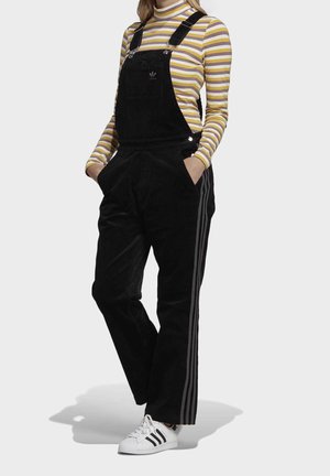 Schwarzer Cord-Latzhose mit verstellbaren Trägern, Seitentaschen und Drei-Streifen-Akzenten. Kombiniert mit einem gestreiften Langarmshirt und weißen Sneakers.