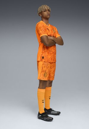 Kit deportivo naranja con un patrón de mármol, acentos negros y un logo. Incluye pantalones cortos a juego, calcetas hasta la rodilla y botas de fútbol negras.