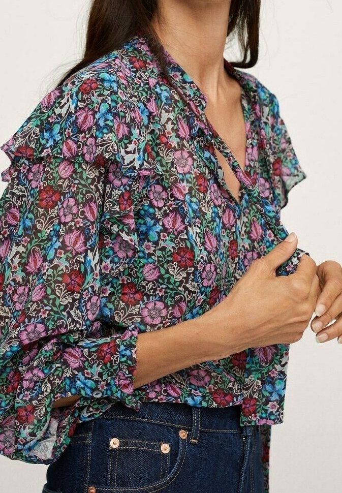 Femme portant un chemisier floral transparent à manches à volants de couleurs violettes, bleues et rouges, associé à un jean en denim bleu foncé.