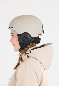 Skihelm in mattem Beige mit verstellbaren schwarzen Ohrpolstern und Belüftungsöffnungen. Tragen mit einer hellbeigen Jacke mit Kapuze.