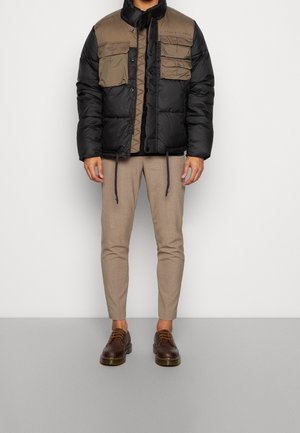Chaqueta acolchada en negro y caqui con cuatro bolsillos frontales, un cuello alto y cordones ajustables; combinada con pantalones beiges y zapatos marrones.
