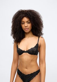 Melna mežģīņu apakšveļas komplekts ar krūšturi ar stīpiņu un ziedu dizainu, komplektā ar atbilstošām mežģīņu biksītēm, izceļot smalku detaļas.