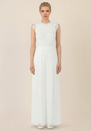 Woman standing straight wearing a sleeveless white lace top and high-waisted wide-leg white trousers against a neutral background.