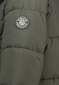 Oliven grüner Pufferjacke mit gestepptem Muster. Verfügt über ein rundes schwarzes Logo-Patch mit der Aufschrift "SUBLEVEL STUDIO COLLECTION" am Ärmel.