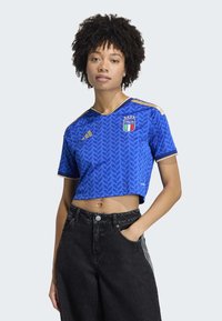 Blue cropped jersey featuring leaf pattern, gold accents, and Italia logo. Paired with high-waisted black jeans. Short sleeves and V-neck design.