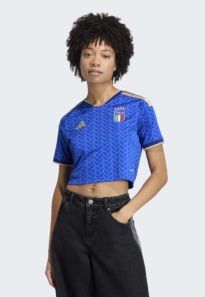 Blue cropped jersey featuring leaf pattern, gold accents, and Italia logo. Paired with high-waisted black jeans. Short sleeves and V-neck design.