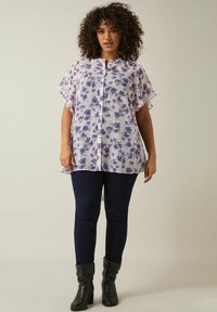 Weißes Blumenblusenoberteil mit lila Blumen, kurze Ärmel, Knopfleiste, kombiniert mit dunkelblauen Skinny Jeans und schwarzen Stiefeletten.