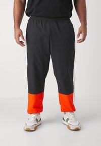 Svarta träningsbyxor med elastisk midja, med reflexdetaljer på sidorna och en orange nederdel, bärs tillsammans med vita sneakers.