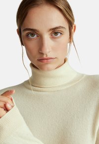 Maglione a collo alto color crema realizzato in un morbido tessuto testurizzato con dettagli a coste sul colletto e sui polsini. Visione ravvicinata del collo e delle maniche.