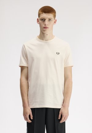 Junger Mann mit kurzen roten Haaren, der ein schlichtes hellbeiges T-Shirt mit einem kleinen schwarzen Lorbeer-Logo auf der Brust trägt, vor einem weißen Hintergrund stehend.