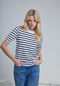 Femme blonde aux longs cheveux portant un t-shirt à manches courtes blanc et bleu marine à rayures et un jean bleu, les mains dans les poches, regardant vers le bas.