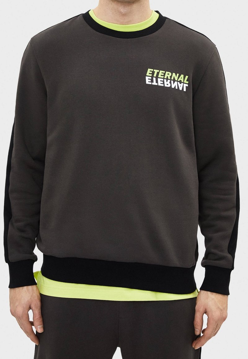 Homme portant un sweat-shirt gris foncé avec des poignets noirs et des accents vert néon, avec « ETERNAL » imprimé deux fois sur la poitrine en vert et blanc.