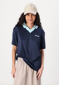 Marinblå kortärmad polo skjorta med vertikala ränder och ljusblå krage. Beigekappa med logodetalj. Bärs med en ljusbeige elastisk kjol.