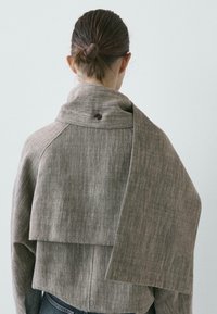 Donna con capelli castani raccolti in uno chignon, indossa una giacca grigia texturizzata con un collo ampio e un pannello di tessuto lungo attaccato, vista da dietro.