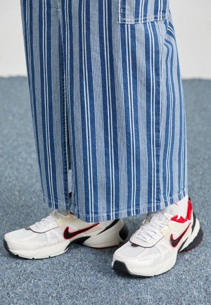 Bílé a červené tenisky Nike nošené s širokými modrobílými pruhovanými kalhotami na texturované modré podlaze.