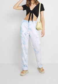 Černý krácený top s uzlovým detailem, světle modré tie-dye cargo kalhoty a béžové platformové boty se žlutými popruhy. Žlutá crossbody taška.