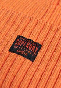 Berretto in maglia a coste arancione con etichetta rettangolare nera che mostra la scritta "SUPERDRY HEAVY DUTY" in testo rosso, caratterizzato da un tessuto testurizzato.