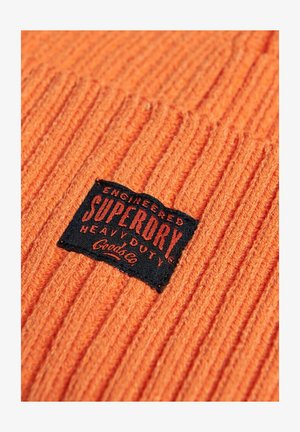 Bonnet en tricot côtelé orange avec une étiquette rectangulaire noire affichant "SUPERDRY HEAVY DUTY" en texte rouge, doté d'un tissu texturé.
