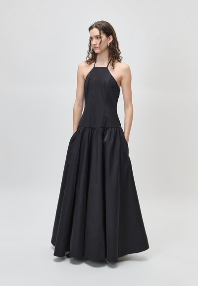 Γυναίκα με σγουρά μαλλιά, ντυμένη με ένα αμάνικο μαύρο φόρεμα halter-neck με φούστα σε πλήρη γραμμή, στέκεται μπροστά σε ένα απλό φωτεινό φόντο.