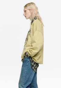 Oluņzaļš lielizmēra krekls ar grafisko dizainu, virsū uz melnas un zaļas rūtainas krekla, kopā ar gaiši ziliem džinsiem.