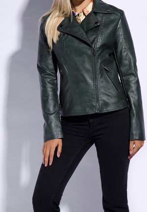 Kvinde iført mørkegrøn læderjakke med asymmetrisk lynlås og sorte bukser, stående med hænder afslappet ved siderne.