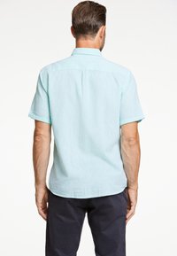 Koszula z krótkim rękawem w jasnym odcieniu teal, wykonana z teksturowanej tkaniny; posiada karczek z tyłu oraz delikatnie zaokrąglony dół, w zestawie z granatowymi spodniami.