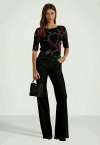 Γυναίκα με μαύρη μπλούζα με σχέδιο αλυσίδας και μαύρα παντελόνια ψηλής μέσης, κρατώντας μια μικρή μαύρη τσάντα, standing against a plain background.