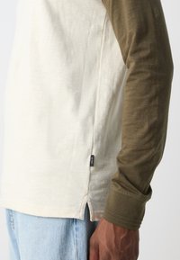 Pikend varrukatega särk, millel on kreemjas keha ja oliivrohelised varrukad, külgedel on lõhikud ning küljepeal väike brändi silt.