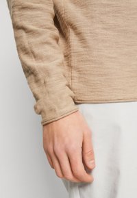 Långärmad, beige topp med en texturerad väv, som har samlademanschetter och en lätt fåll. Hand som vilar på ljusfärgad tyg.