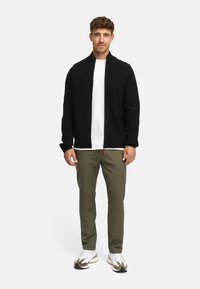 Zwarte gebreide cardigan met hoge kraag, gedragen over een witte t-shirt. Olijfgroene broek en witte sneakers met grijze en zwarte accenten zijn ook zichtbaar.