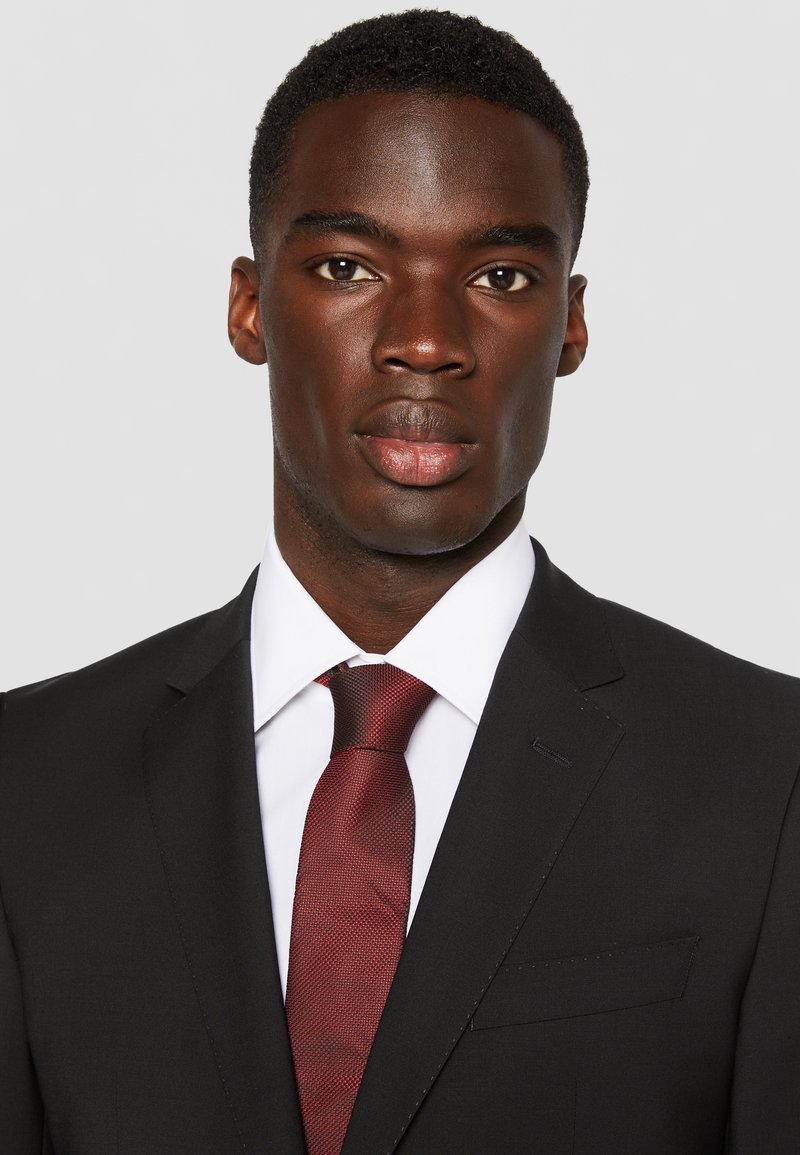 Zwarte pak met een op maat gemaakte pasvorm, wit overhemd en een gestructureerde rode stropdas. De outfit heeft een spitse revers en minimalistische ontwerpelementen.