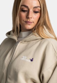 Sudadera con cremallera beige, hecha de una tela suave, con un logo en la parte frontal y un pequeño bordado de mariposa morada en el lado izquierdo del pecho.