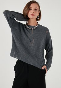 Dunkelgrauer, locker sitzender Rippstrickpullover mit Rundhalsausschnitt und distressed Details. Kombiniert mit schwarzen Hosen und einer klobigen Perlenkette.