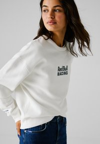 Sudadera blanca con el logo "Red Bull RACING" en azul oscuro. Presenta un cuello redondo y mangas con puños. Combinada con jeans de mezclilla azul.