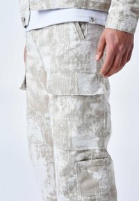 Pantalones cargo en beige y blanco con un patrón abstracto texturizado. Cuenta con bolsillos laterales y una etiqueta de logo sutil en la cinturilla.