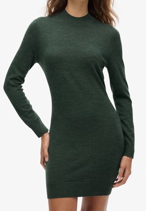 Kvinde iført en tætsiddende, mørkegrøn, langærmet sweaterkjole med rund halsudskæring, stående med den ene hånd på hoften.