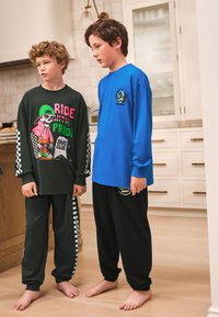 Deux garçons portant des vêtements de détente assortis. L'un porte un t-shirt noir à manches longues avec un graphisme coloré et des accents verts, associé à un pantalon sombre à motif à carreaux. L'autre porte un t-shirt bleu à manches longues et un pantalon de survêtement noir avec un logo. Tous deux sont pieds nus.